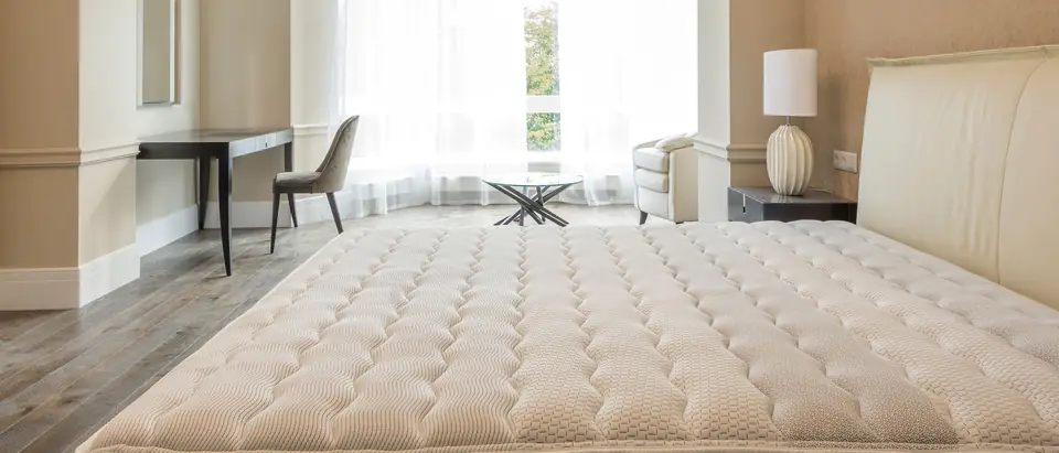 A bare quilted mattress on a bed in a bright bedroom with a bay window and a desk
