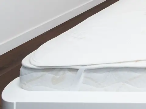The corner of a white mattress with a corded piped seam along its top edge, under a mattress pad held by an elastic strap