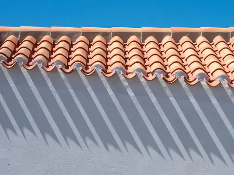 The edge of a clay barrel tile roof overhanging a white stucco wall, with the tiles casting a row of pointed shadows